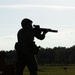 A silhouetted figure in full tactical gear is aiming a rifle equipped with a suppressor and optic sight at an outdoor shooting range. The backlit composition renders the subject entirely in shadow against a bright sky and open grassy field. The setting appears to be a military or law enforcement training range based on the infrastructure and equipment visible.