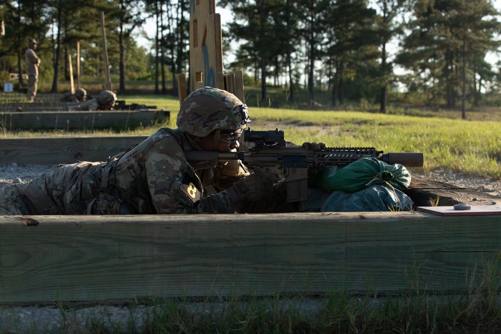 A U.S. Army soldier in full combat gear is positioned in a prone firing position behind a wooden barrier at what appears to be a military rifle range. The soldier is aiming a rifle with optical sights, using a sandbag for weapon support. Additional soldiers and range infrastructure are visible in the background.