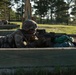 A U.S. Army soldier in full combat gear is positioned in a prone firing position behind a wooden barrier at what appears to be a military rifle range. The soldier is aiming a rifle with optical sights, using a sandbag for weapon support. Additional soldiers and range infrastructure are visible in the background.