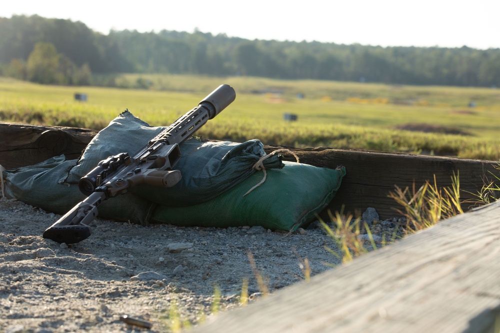 A scoped rifle with a suppressor attached is resting on green sandbags behind a wooden barrier, consistent with a shooting range firing position. The weapon appears to be a modern semi-automatic rifle with a bipod deployed and an optical scope mounted. The setting suggests a military or civilian long-range shooting range with an open field downrange.