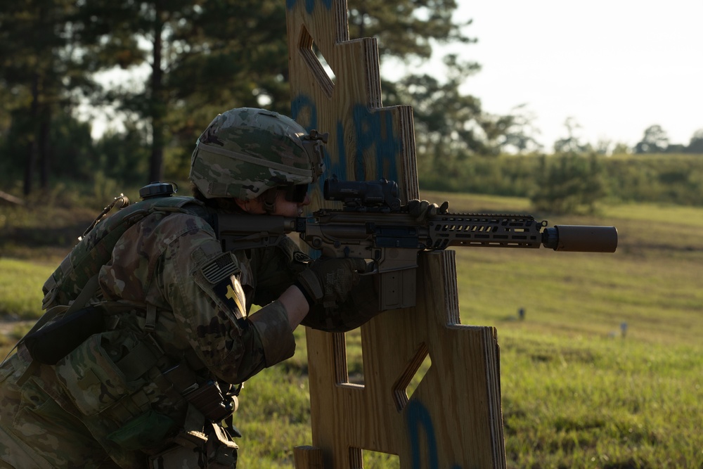 A U.S. Army soldier in full combat gear is taking an aiming position behind a wooden barricade structure on a training range. The soldier is equipped with a suppressed rifle, tactical vest, helmet, and multicam camouflage uniform. The setting appears to be an outdoor military training facility with open fields and trees in the background.