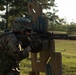 A U.S. Army soldier in full combat gear is taking an aiming position behind a wooden barricade structure on a training range. The soldier is equipped with a suppressed rifle, tactical vest, helmet, and multicam camouflage uniform. The setting appears to be an outdoor military training facility with open fields and trees in the background.