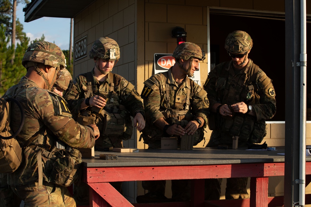 Four soldiers in US Army OCP (Operational Camouflage Pattern) uniforms are gathered around a red-framed outdoor table at what appears to be a military training range or station, building number 8528. They appear to be handling ammunition or small equipment items, likely during a pre-range briefing or ammunition draw. The setting suggests a training exercise at a military installation, possibly Fort Bragg or a similar US Army post.