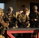 Four soldiers in US Army OCP (Operational Camouflage Pattern) uniforms are gathered around a red-framed outdoor table at what appears to be a military training range or station, building number 8528. They appear to be handling ammunition or small equipment items, likely during a pre-range briefing or ammunition draw. The setting suggests a training exercise at a military installation, possibly Fort Bragg or a similar US Army post.