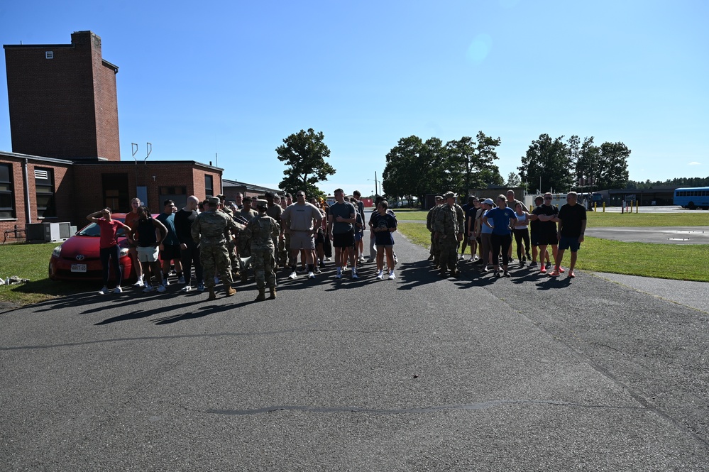 A large group of individuals, including uniformed military personnel and civilians in athletic wear, are gathered on a paved area at what appears to be a military installation. The group appears to be assembling or receiving instructions, possibly before or after a physical training event. The setting includes brick buildings, trees, and a blue bus visible in the background.