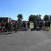 A large group of individuals, including uniformed military personnel and civilians in athletic wear, are gathered on a paved area at what appears to be a military installation. The group appears to be assembling or receiving instructions, possibly before or after a physical training event. The setting includes brick buildings, trees, and a blue bus visible in the background.