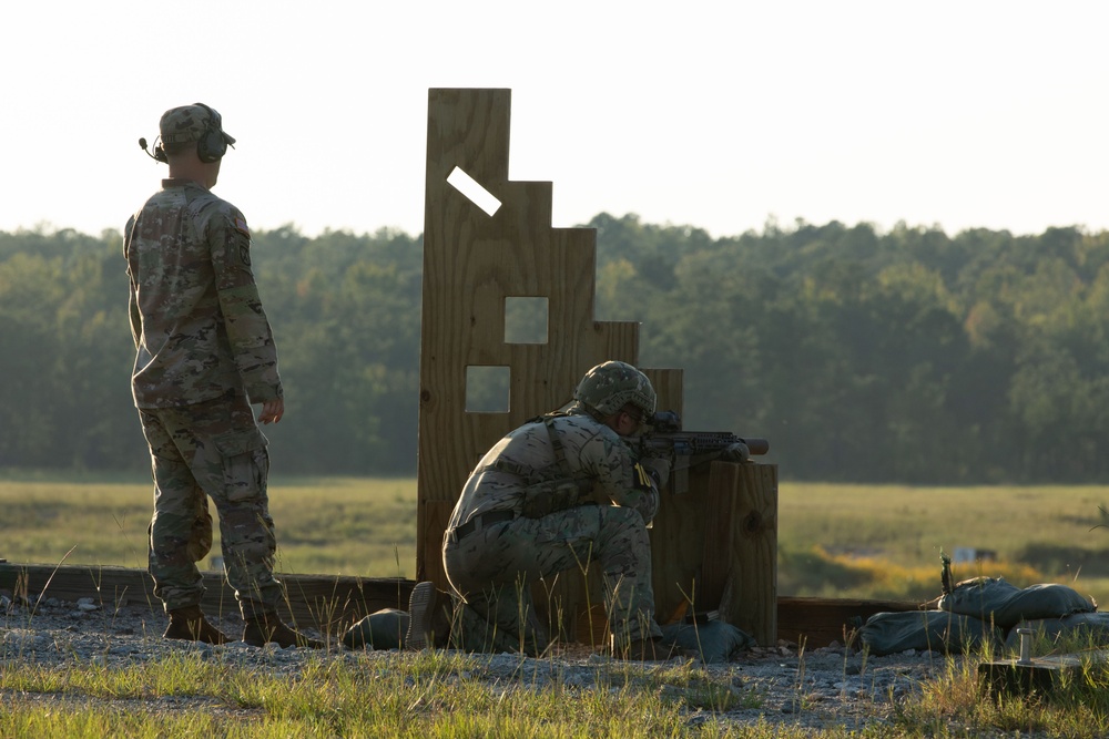 Two military personnel are on an outdoor shooting range during what appears to be early morning or late afternoon based on the low-angle sunlight. One soldier stands observing while a second soldier crouches in a firing position behind a wooden barricade structure, aiming a rifle downrange. The setting appears to be a military training facility with an open field and tree line in the background.