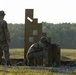 Two military personnel are on an outdoor shooting range during what appears to be early morning or late afternoon based on the low-angle sunlight. One soldier stands observing while a second soldier crouches in a firing position behind a wooden barricade structure, aiming a rifle downrange. The setting appears to be a military training facility with an open field and tree line in the background.