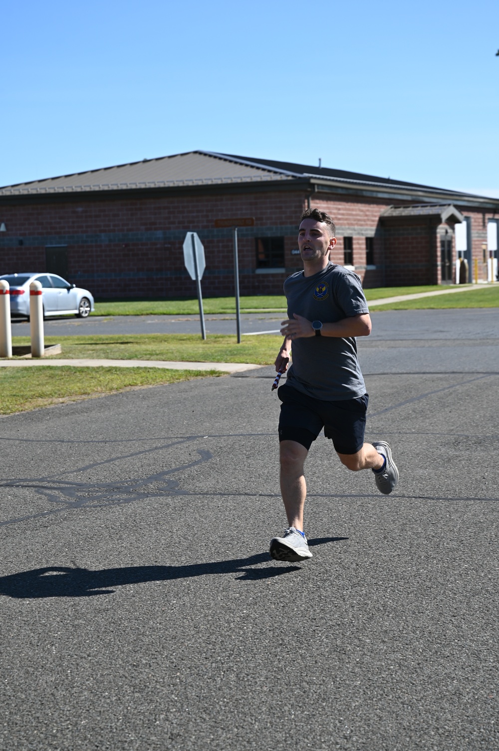 A young man is running on a paved road in what appears to be a military base or institutional campus setting. He is wearing athletic clothing including a gray t-shirt with a military-style emblem, dark shorts, and gray running shoes. The background features a red brick building, manicured grass, a stop sign, and a parked white sedan under a clear blue sky.