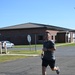A young man is running on a paved road in what appears to be a military base or institutional campus setting. He is wearing athletic clothing including a gray t-shirt with a military-style emblem, dark shorts, and gray running shoes. The background features a red brick building, manicured grass, a stop sign, and a parked white sedan under a clear blue sky.