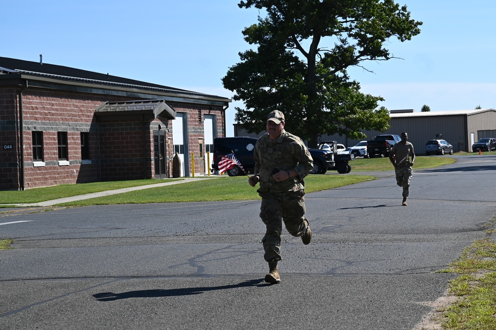 Two military personnel in camouflage uniforms are running along a paved road on what appears to be a military installation or base. The lead runner is carrying a small American flag and wearing a cap. The setting includes brick buildings, parked vehicles, and a large tree under clear blue skies.