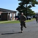 Two military personnel in camouflage uniforms are running along a paved road on what appears to be a military installation or base. The lead runner is carrying a small American flag and wearing a cap. The setting includes brick buildings, parked vehicles, and a large tree under clear blue skies.