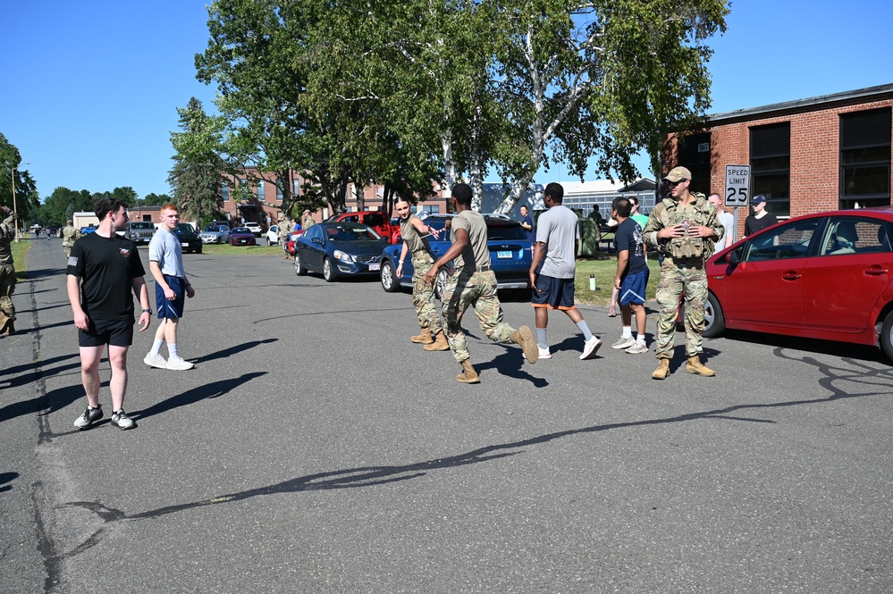 The image shows a group of military personnel in camouflage uniforms interacting with civilians in athletic wear in a parking lot on what appears to be a military installation. The setting includes brick buildings, parked cars, and a speed limit sign showing 25 mph. The scene appears to be an informal gathering or orientation activity during daytime under clear blue skies.