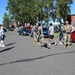 The image shows a group of military personnel in camouflage uniforms interacting with civilians in athletic wear in a parking lot on what appears to be a military installation. The setting includes brick buildings, parked cars, and a speed limit sign showing 25 mph. The scene appears to be an informal gathering or orientation activity during daytime under clear blue skies.