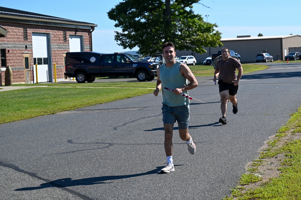 Two men are running on a paved road near what appears to be a military or government facility. The lead runner, wearing a teal tank top and shorts, is smiling and holding a red-and-white striped baton or finish tape, suggesting he is winning a race. A second runner in a brown shirt follows behind at a distance.