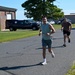 Two men are running on a paved road near what appears to be a military or government facility. The lead runner, wearing a teal tank top and shorts, is smiling and holding a red-and-white striped baton or finish tape, suggesting he is winning a race. A second runner in a brown shirt follows behind at a distance.