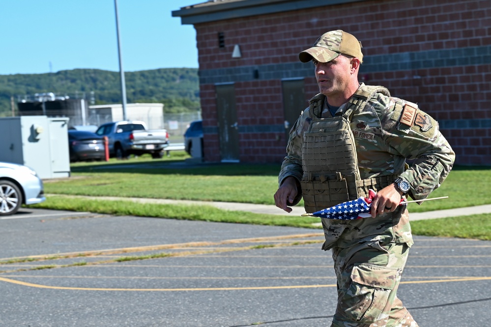 A U.S. Air Force service member in full OCP (Operational Camouflage Pattern) uniform and plate carrier vest is walking or jogging across a paved area on what appears to be a military installation. He is carrying a folded American flag and wearing a tan baseball cap. The setting appears to be a sunny day at a base facility with brick buildings and parked vehicles in the background.