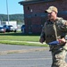 A U.S. Air Force service member in full OCP (Operational Camouflage Pattern) uniform and plate carrier vest is walking or jogging across a paved area on what appears to be a military installation. He is carrying a folded American flag and wearing a tan baseball cap. The setting appears to be a sunny day at a base facility with brick buildings and parked vehicles in the background.