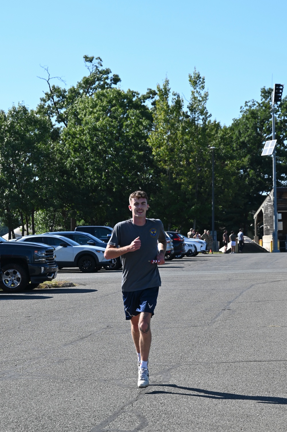 A young man is running through a paved parking lot area on a clear, sunny day. He is wearing athletic clothing including a gray t-shirt with a military-style patch/emblem, navy blue shorts with what appears to be a US Air Force logo, and white sneakers. People and parked vehicles are visible in the background.