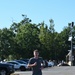 A young man is running through a paved parking lot area on a clear, sunny day. He is wearing athletic clothing including a gray t-shirt with a military-style patch/emblem, navy blue shorts with what appears to be a US Air Force logo, and white sneakers. People and parked vehicles are visible in the background.