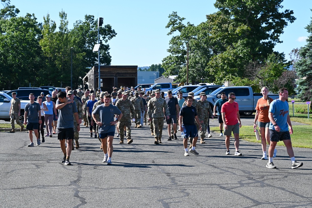 A large group of military personnel and civilians are walking across a paved parking lot area, likely following a physical training event or formation activity. The group includes individuals in military camouflage uniforms (ACU/OCP pattern) mixed with civilians in casual athletic wear. The setting appears to be a military installation or base during daytime in warm weather.