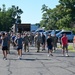 A large group of military personnel and civilians are walking across a paved parking lot area, likely following a physical training event or formation activity. The group includes individuals in military camouflage uniforms (ACU/OCP pattern) mixed with civilians in casual athletic wear. The setting appears to be a military installation or base during daytime in warm weather.