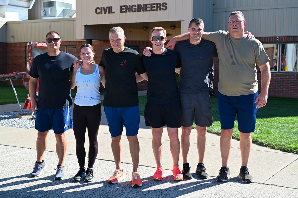 Six individuals pose together in a group photo in front of a building labeled 'Civil Engineers.' They are dressed in athletic/workout attire including shorts, t-shirts, and running shoes, suggesting they have completed or are about to begin a physical fitness activity. The setting appears to be a military or government facility based on the building's architecture and signage.