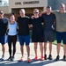 Six individuals pose together in a group photo in front of a building labeled 'Civil Engineers.' They are dressed in athletic/workout attire including shorts, t-shirts, and running shoes, suggesting they have completed or are about to begin a physical fitness activity. The setting appears to be a military or government facility based on the building's architecture and signage.
