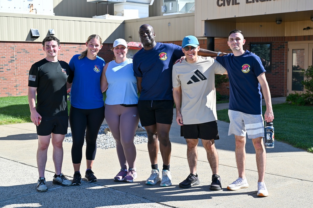 A group of seven people pose together outdoors in athletic/workout attire in front of a brick building with a 'Civil Engi...' sign visible. Several members are wearing matching navy blue shirts with what appears to be a unit or squadron patch. The setting and attire suggest a military or government facility fitness event or group workout.