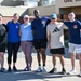 A group of seven people pose together outdoors in athletic/workout attire in front of a brick building with a 'Civil Engi...' sign visible. Several members are wearing matching navy blue shirts with what appears to be a unit or squadron patch. The setting and attire suggest a military or government facility fitness event or group workout.