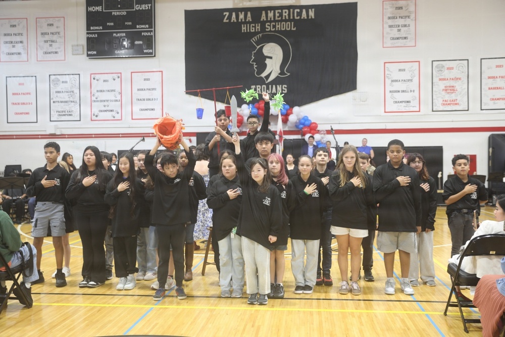 A group of students dressed in matching black sweatshirts stand in a school gymnasium at Zama American High School, with most placing their right hands over their hearts, suggesting they are reciting the Pledge of Allegiance or a similar patriotic ceremony. Some students in the back row are holding colorful props including what appears to be a piñata, a blue bucket, and decorative items, indicating a multicultural or festive school event. The setting is a decorated gymnasium with championship banners, a Trojans mascot banner, and red, white, and blue balloons.
