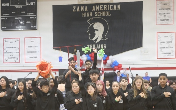 A group of students dressed in matching black sweatshirts stand in a school gymnasium at Zama American High School, with most placing their right hands over their hearts, suggesting they are reciting the Pledge of Allegiance or a similar patriotic ceremony. Some students in the back row are holding colorful props including what appears to be a piñata, a blue bucket, and decorative items, indicating a multicultural or festive school event. The setting is a decorated gymnasium with championship banners, a Trojans mascot banner, and red, white, and blue balloons.