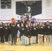 A group of students dressed in matching black sweatshirts stand in a school gymnasium at Zama American High School, with most placing their right hands over their hearts, suggesting they are reciting the Pledge of Allegiance or a similar patriotic ceremony. Some students in the back row are holding colorful props including what appears to be a piñata, a blue bucket, and decorative items, indicating a multicultural or festive school event. The setting is a decorated gymnasium with championship banners, a Trojans mascot banner, and red, white, and blue balloons.