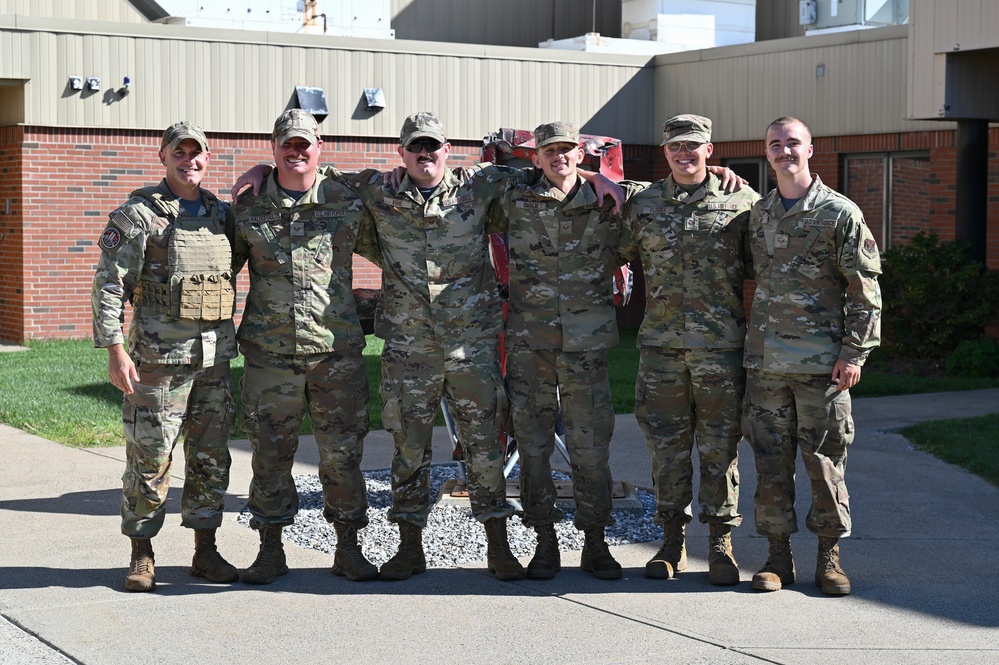 Six military personnel in OCP (Operational Camouflage Pattern) uniforms pose together outdoors in a group photo. They are standing in a row with arms around each other's shoulders in a relaxed, informal manner. The setting appears to be a military installation with brick buildings visible in the background.