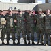 Six military personnel in OCP (Operational Camouflage Pattern) uniforms pose together outdoors in a group photo. They are standing in a row with arms around each other's shoulders in a relaxed, informal manner. The setting appears to be a military installation with brick buildings visible in the background.