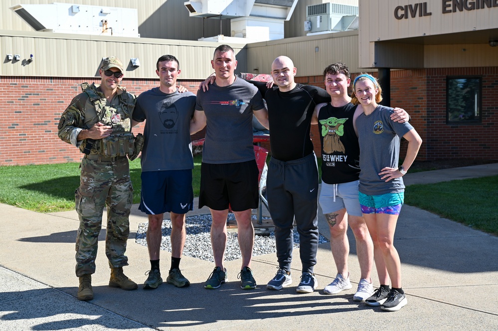 Six individuals pose for a group photo outside a building marked 'Civil Engi[neering]', likely on a military installation. One person on the left is wearing full camouflage military uniform with tactical vest, while the remaining five are dressed in casual athletic/workout clothing. The group appears to be gathered after a physical fitness event or workout session.