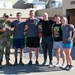 Six individuals pose for a group photo outside a building marked 'Civil Engi[neering]', likely on a military installation. One person on the left is wearing full camouflage military uniform with tactical vest, while the remaining five are dressed in casual athletic/workout clothing. The group appears to be gathered after a physical fitness event or workout session.