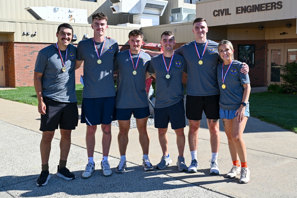 Six individuals wearing matching gray t-shirts with unit patches are posing together in front of a building labeled 'Civil Engineers.' All six are wearing gold medals on red, white, and blue ribbons, suggesting they recently competed in and won a sporting or fitness event. The group appears to be affiliated with a military or civil engineering unit.