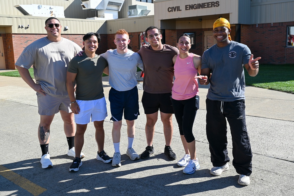 Six individuals in athletic/casual workout attire pose together in front of a building labeled 'Civil Engineers,' likely a military or government facility. The group appears to be in good spirits after a physical training or fitness activity, smiling and standing arm-in-arm. The setting suggests this may be a military base civil engineering unit.