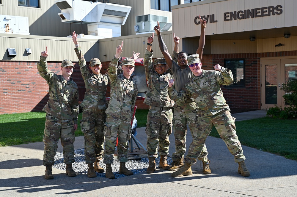 Six military personnel in OCP (Operational Camouflage Pattern) uniforms pose enthusiastically in front of a 'Civil Engineers' building, raising their index fingers in a 'number one' gesture. The group appears celebratory, suggesting a team achievement or milestone. The setting appears to be a military installation, likely an Air Force base given the Civil Engineers signage.