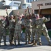 Six military personnel in OCP (Operational Camouflage Pattern) uniforms pose enthusiastically in front of a 'Civil Engineers' building, raising their index fingers in a 'number one' gesture. The group appears celebratory, suggesting a team achievement or milestone. The setting appears to be a military installation, likely an Air Force base given the Civil Engineers signage.