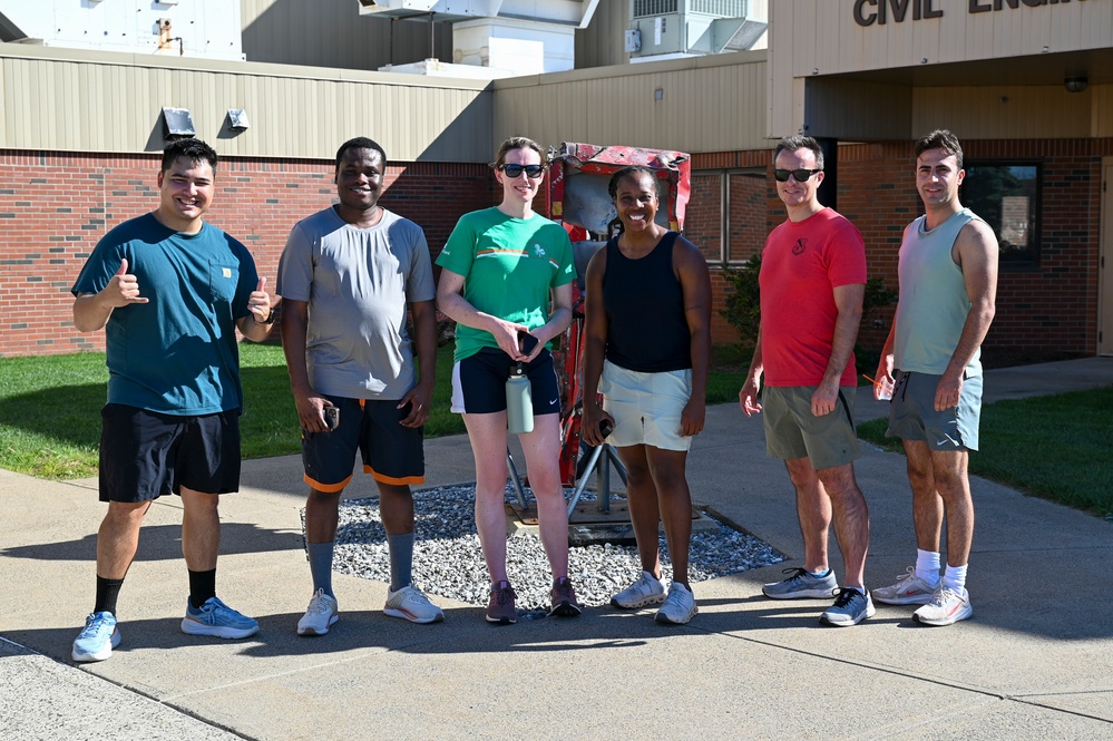 A group of six people pose together outdoors in front of a building partially labeled 'Civil Engi...' (likely Civil Engineering). They are dressed in athletic/casual wear, suggesting a fitness or recreational activity. One person on the left is giving a shaka/thumbs-up gesture, and the group appears to be in good spirits.