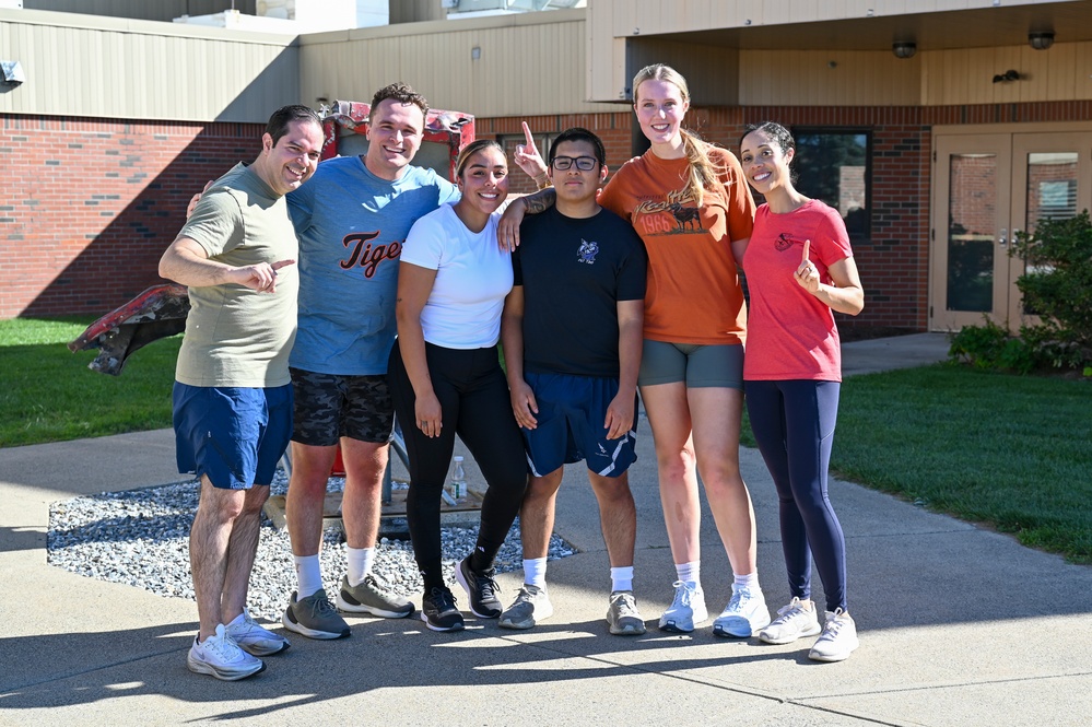 A group of six people pose together outdoors in front of a brick building, smiling for a group photo. They are dressed in casual athletic wear, and several individuals are making 'number one' finger gestures. The setting appears to be a school or institutional campus on a sunny day.