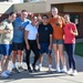 A group of six people pose together outdoors in front of a brick building, smiling for a group photo. They are dressed in casual athletic wear, and several individuals are making 'number one' finger gestures. The setting appears to be a school or institutional campus on a sunny day.