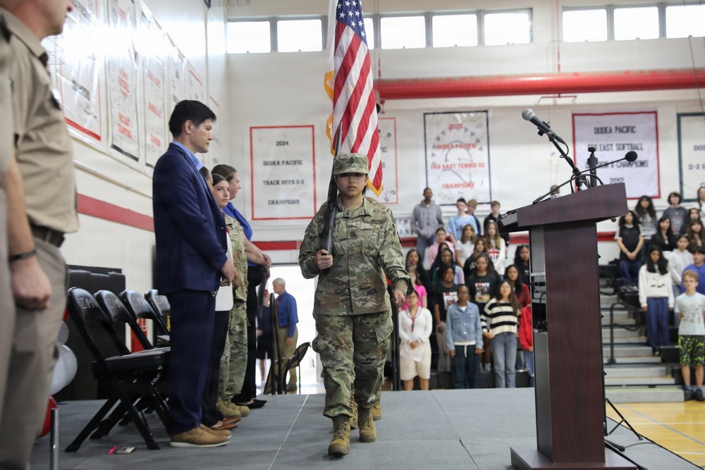 A school assembly or ceremony is taking place in a gymnasium, where a student in military camouflage uniform is carrying the American flag during what appears to be a color guard presentation. Banners on the walls reference DoDEA Pacific championships, indicating this is likely a Department of Defense Dependents Schools facility. Students and adults are standing respectfully in the bleachers and on the gym floor.