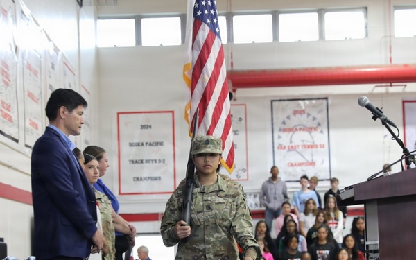 A school assembly or ceremony is taking place in a gymnasium, where a student in military camouflage uniform is carrying the American flag during what appears to be a color guard presentation. Banners on the walls reference DoDEA Pacific championships, indicating this is likely a Department of Defense Dependents Schools facility. Students and adults are standing respectfully in the bleachers and on the gym floor.