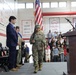 A school assembly or ceremony is taking place in a gymnasium, where a student in military camouflage uniform is carrying the American flag during what appears to be a color guard presentation. Banners on the walls reference DoDEA Pacific championships, indicating this is likely a Department of Defense Dependents Schools facility. Students and adults are standing respectfully in the bleachers and on the gym floor.