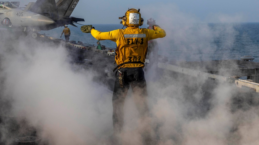A U.S. Navy flight deck crew member in a yellow jersey stands on an aircraft carrier flight deck surrounded by steam, arms outstretched in a directing signal. The individual is identified as belonging to 'FLY-3' division with the number '342' on their back. A military jet aircraft (numbered 405) is visible in the background along with other deck personnel.