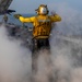 A U.S. Navy flight deck crew member in a yellow jersey stands on an aircraft carrier flight deck surrounded by steam, arms outstretched in a directing signal. The individual is identified as belonging to 'FLY-3' division with the number '342' on their back. A military jet aircraft (numbered 405) is visible in the background along with other deck personnel.