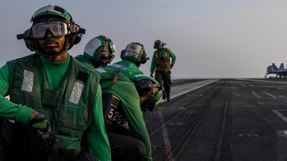 The image shows U.S. Navy flight deck crew members wearing green jerseys, helmets, and protective gear on an aircraft carrier flight deck. Multiple personnel are visible in a line formation, with at least one aircraft visible in the background on the right side. The scene appears to be during active flight deck operations.